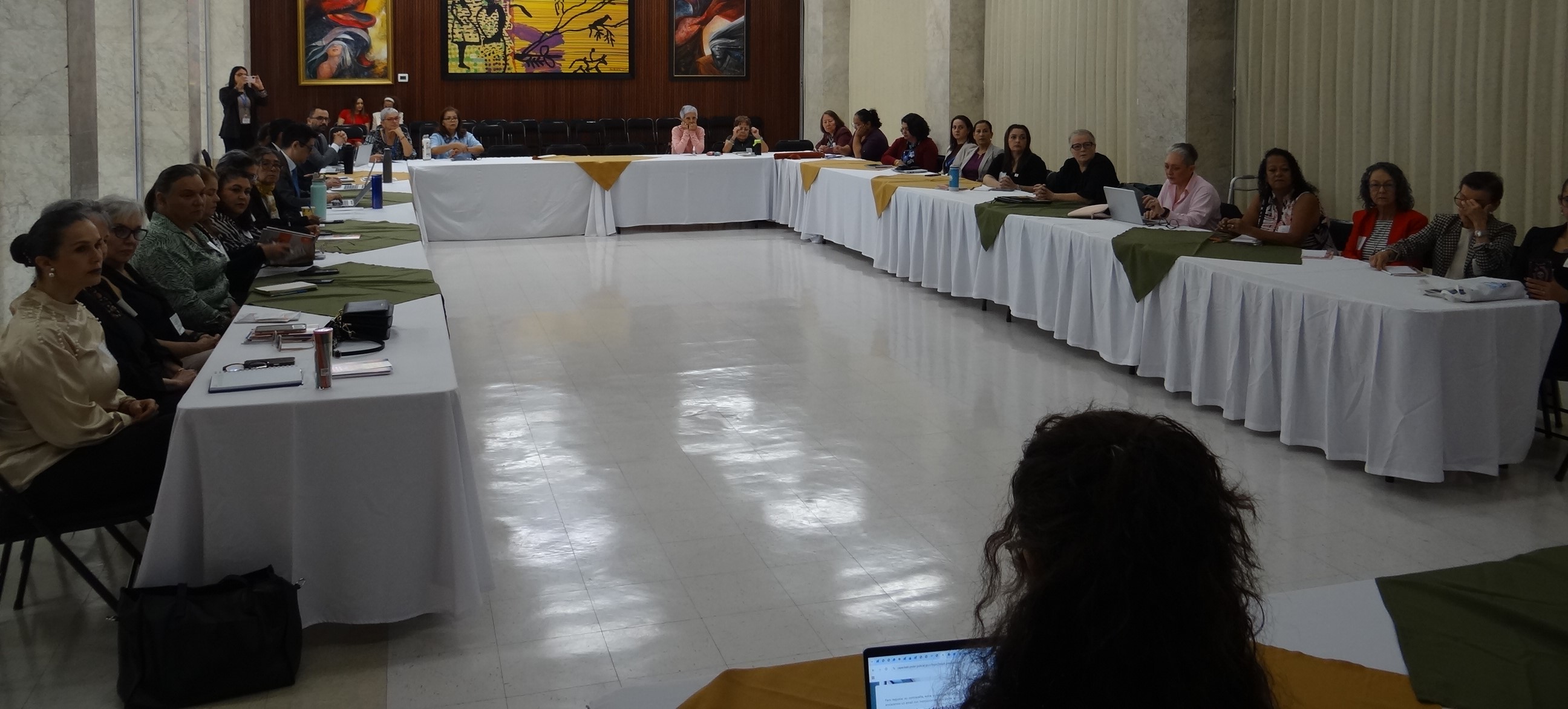 Las mesas están organizadas en forma de "U", cubiertas con manteles blancos y caminos de mesa en tonos verde y dorado. Alrededor de ellas se encuentran numerosas personas asistentes, distribuidas de manera uniforme y orientadas hacia el espacio central de la sala, lo que favorece la interacción y la discusión grupal.